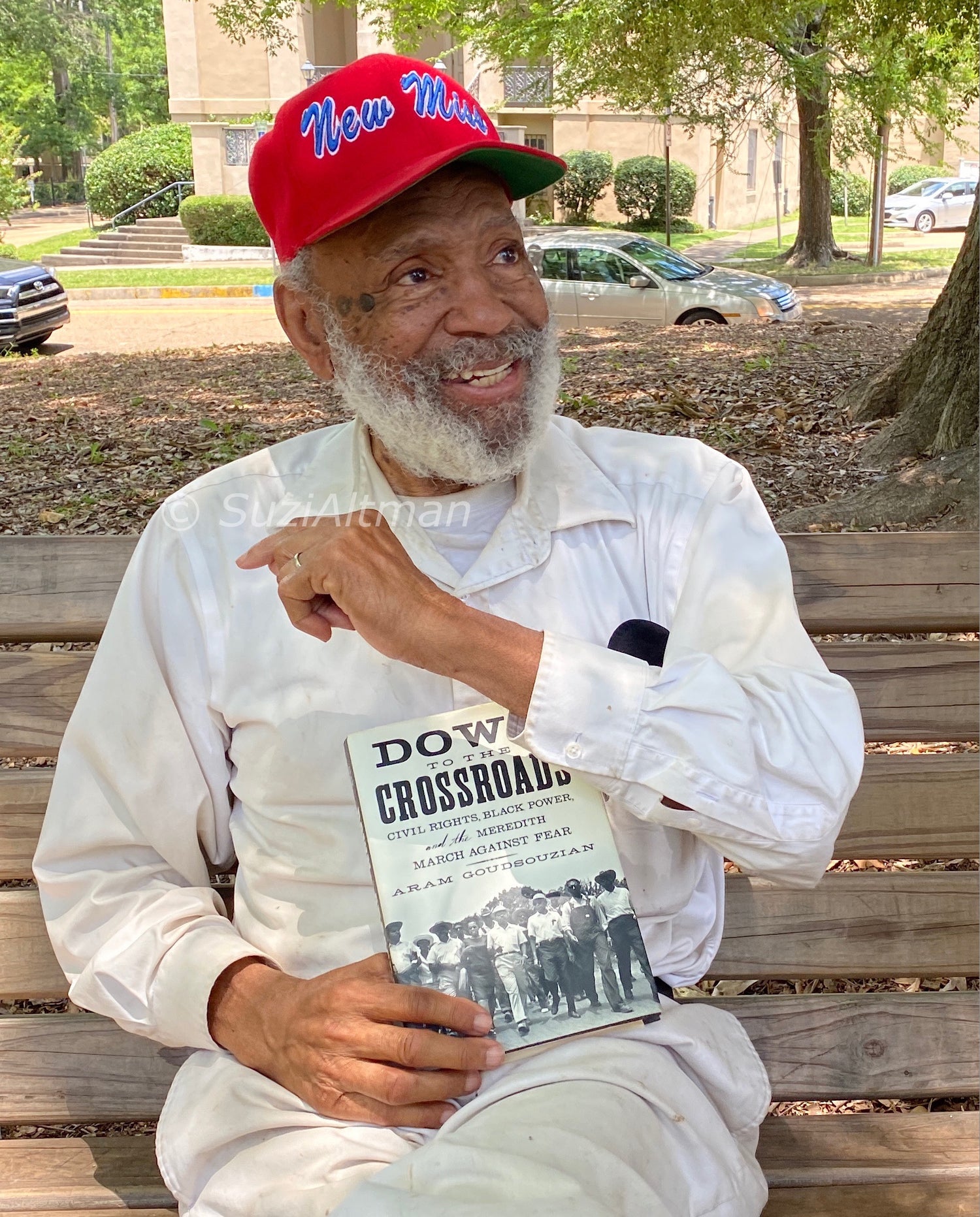 James Meredith smiling on shaded park bench wearing bright red baseball cap with New Miss embroidered in blue script 